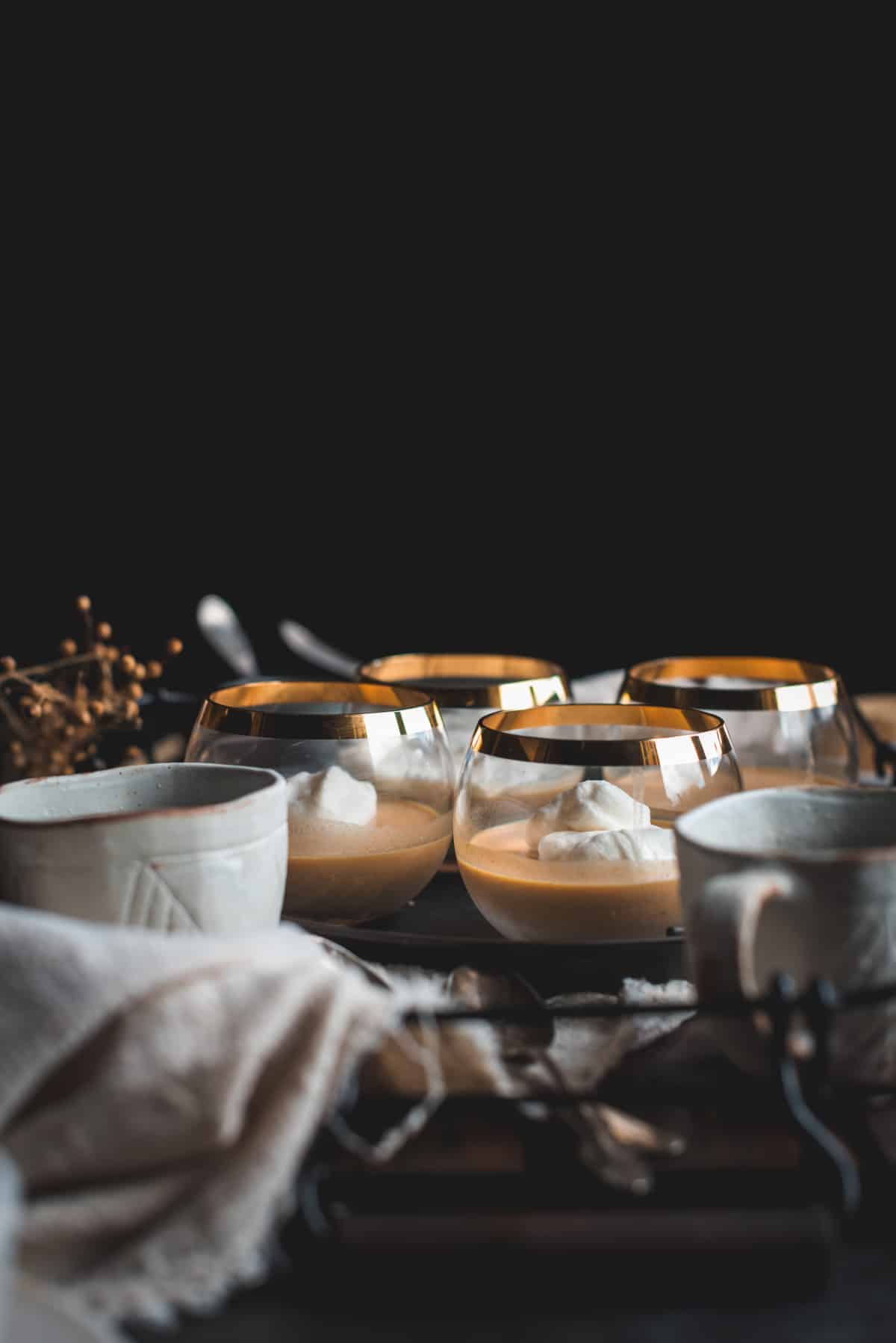 4 small gold rimmed glass bowls are filled with Pumpkin Panna Cotta and topped with whipped cream. There is various other items on the dark colored table like ceramic bowls and jugs, and dried flowers.