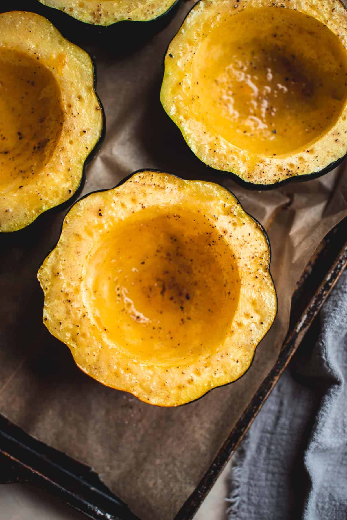 On a lined baking tray, 4 halves of acorn squash have been de-seeded and baked in the oven with olive oil on top.