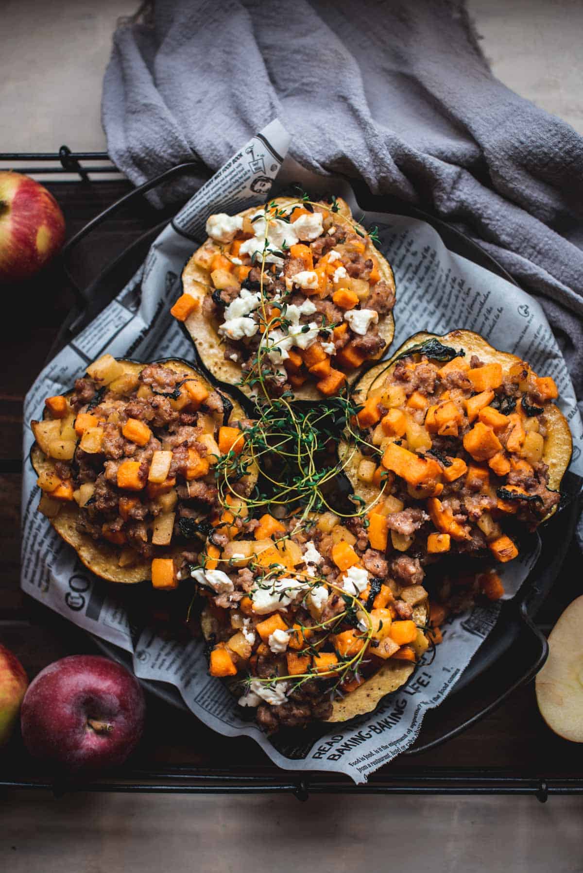 4 half acorn squash stuffed with bright orange ingredients like spiced sausage meat and sweet potato. They are sitting on a newspaper lined plate and have been garnished with some cheese and herbs.