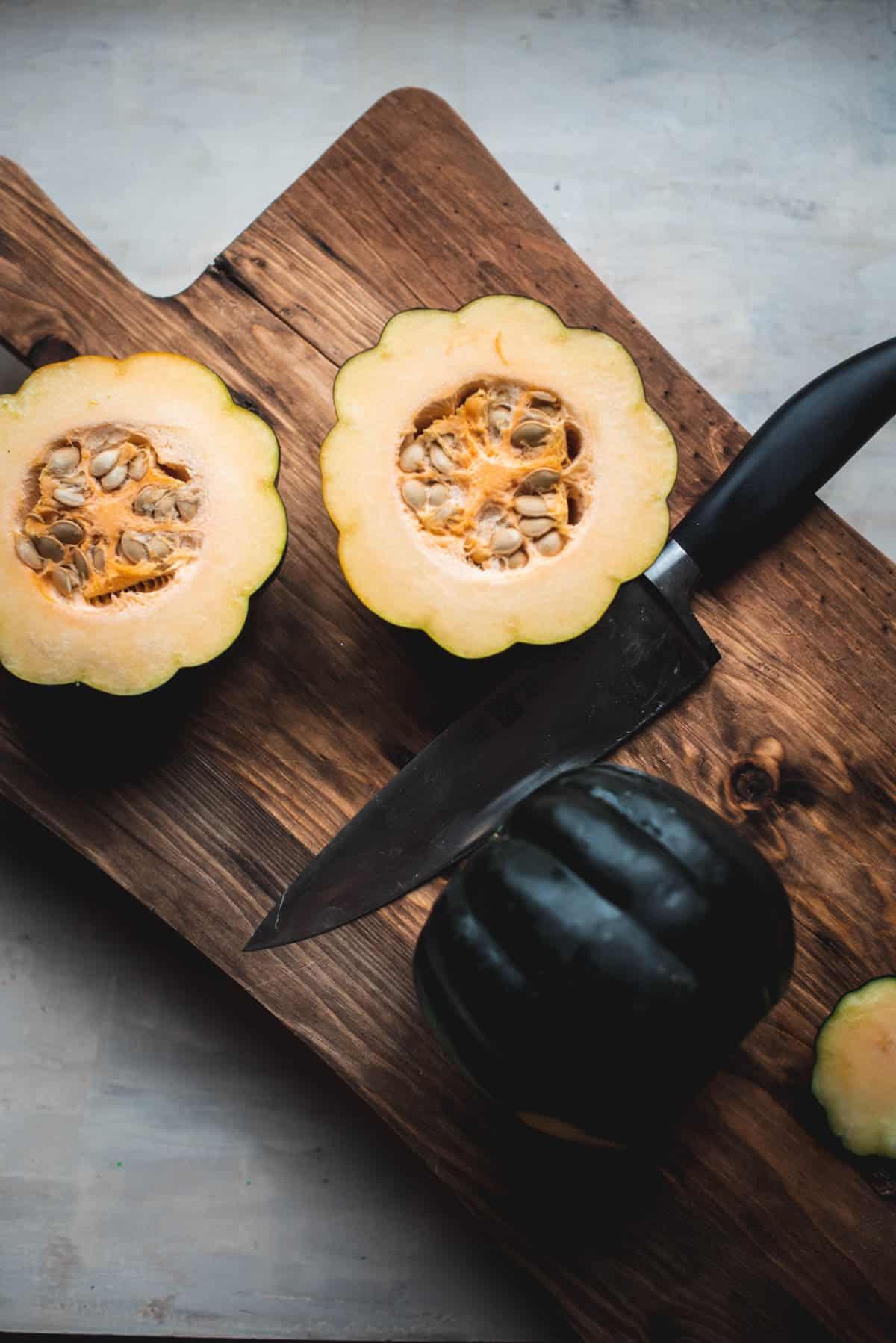 An acorn squash has been cut in half and sitting on a wooden chopping board. The half pieces of squash are still filled with seeds and another squash is whole on the chopping board, ready to be slice by the knife resting next to it.