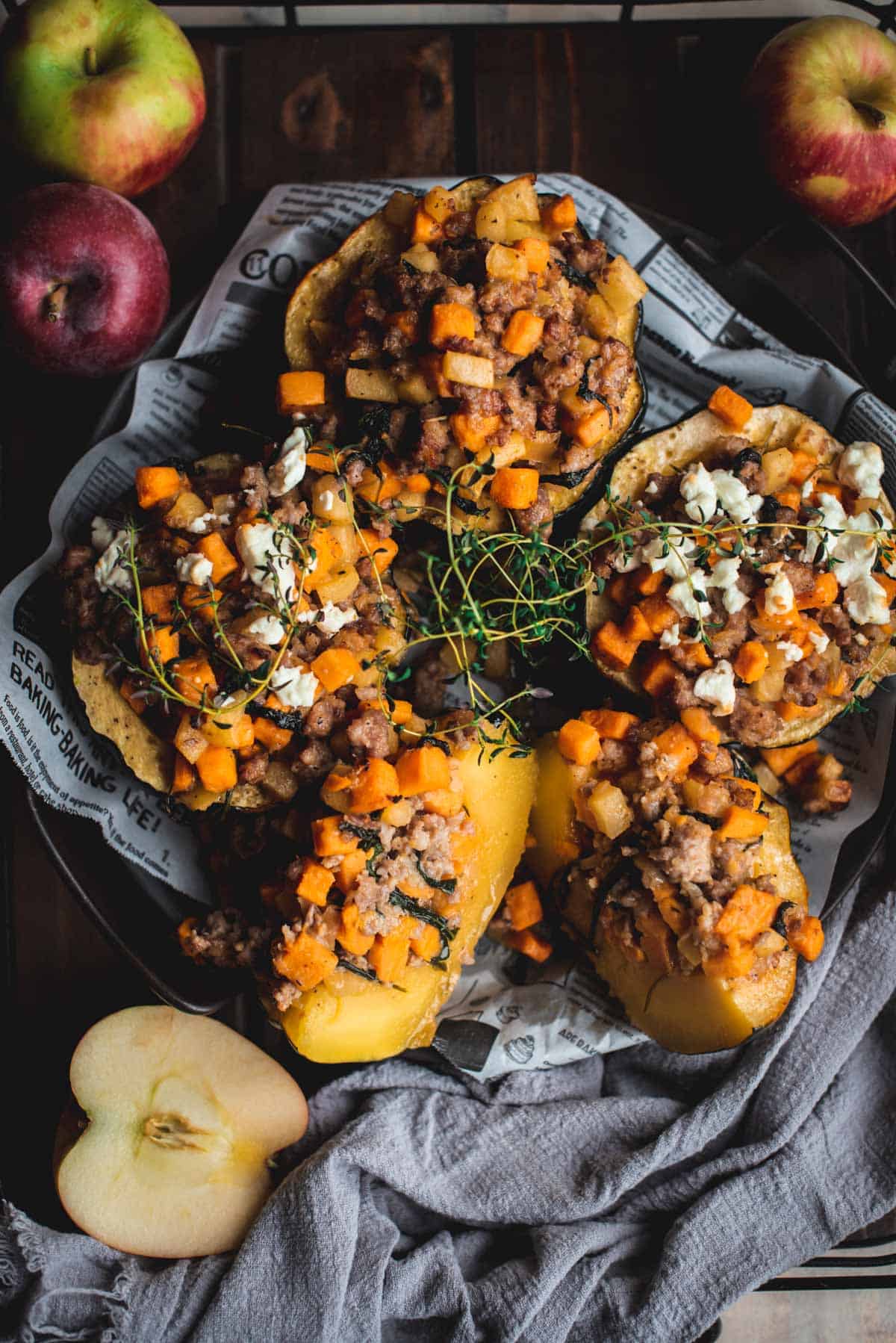 View from above of sausage stuffed acorn squash sitting on a newspaper lined plate. 3 of the half acorns are still intact and 1 half has been sliced in 2.  The color of the dish is very warm with lots of shades of orange are they are topped with green herbs. There is a discarded half apple in the bottom corner resting on a blue dish cloth.