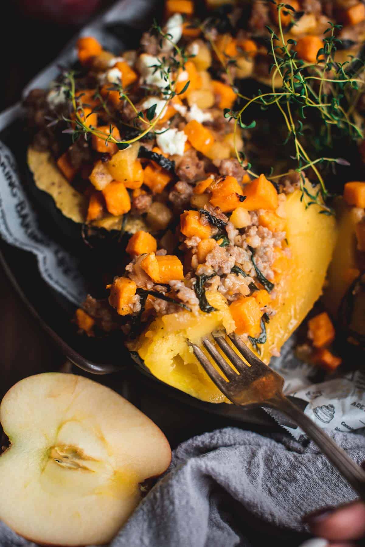 Close up picture of sausage stuffed acorn squash. You can see the acorn squash has been cut into and you can see all the different components to the stuffing like sausage meat, sweet potato, apples, onions and herbs. A fork is inside part of the squash, ready to scoop up another bite. 
An extra half apple is left to the side of plate.