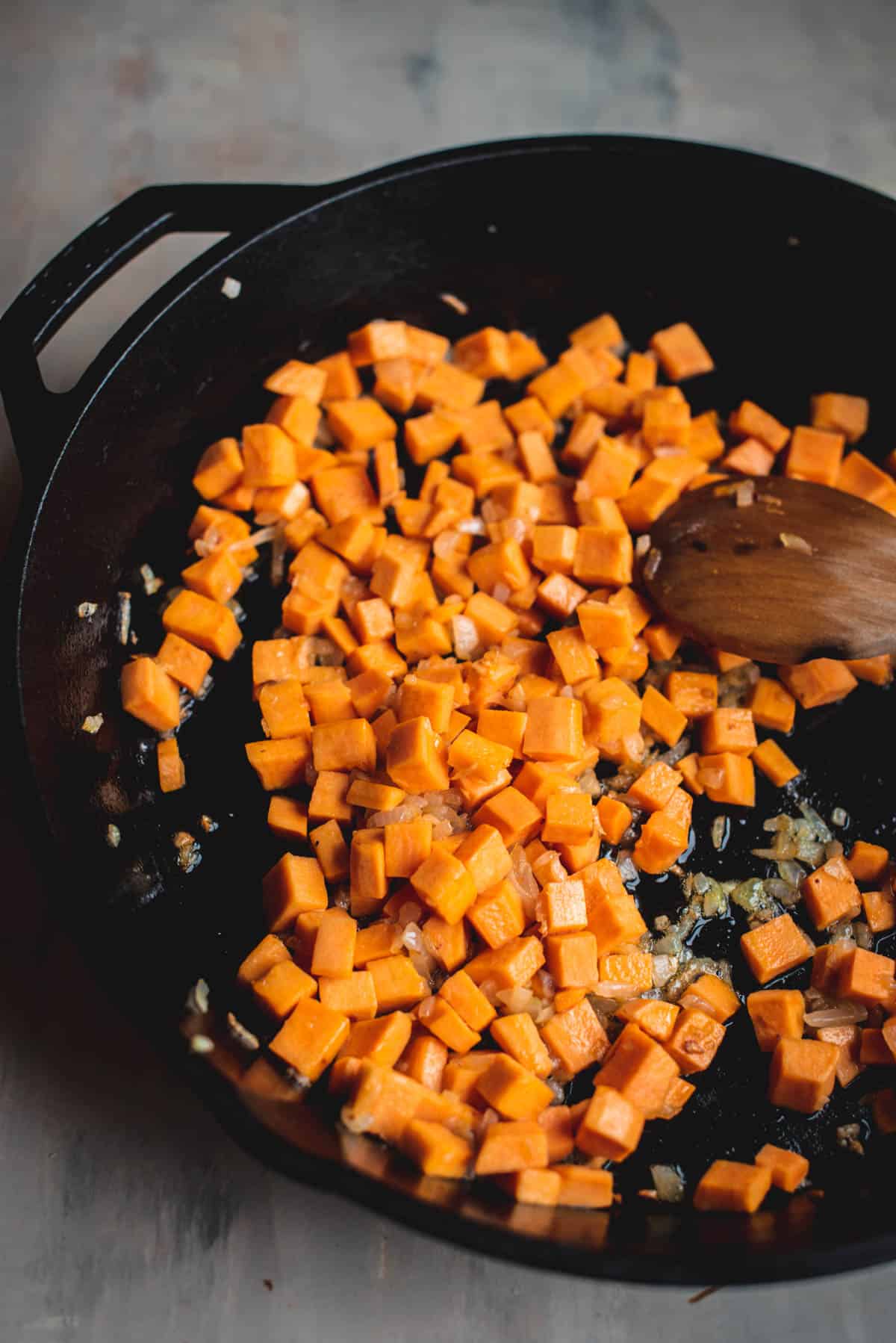 A black skillet is on the oven and is cooking the stuffing mixture, made up of sweet potato, onions and garlic.