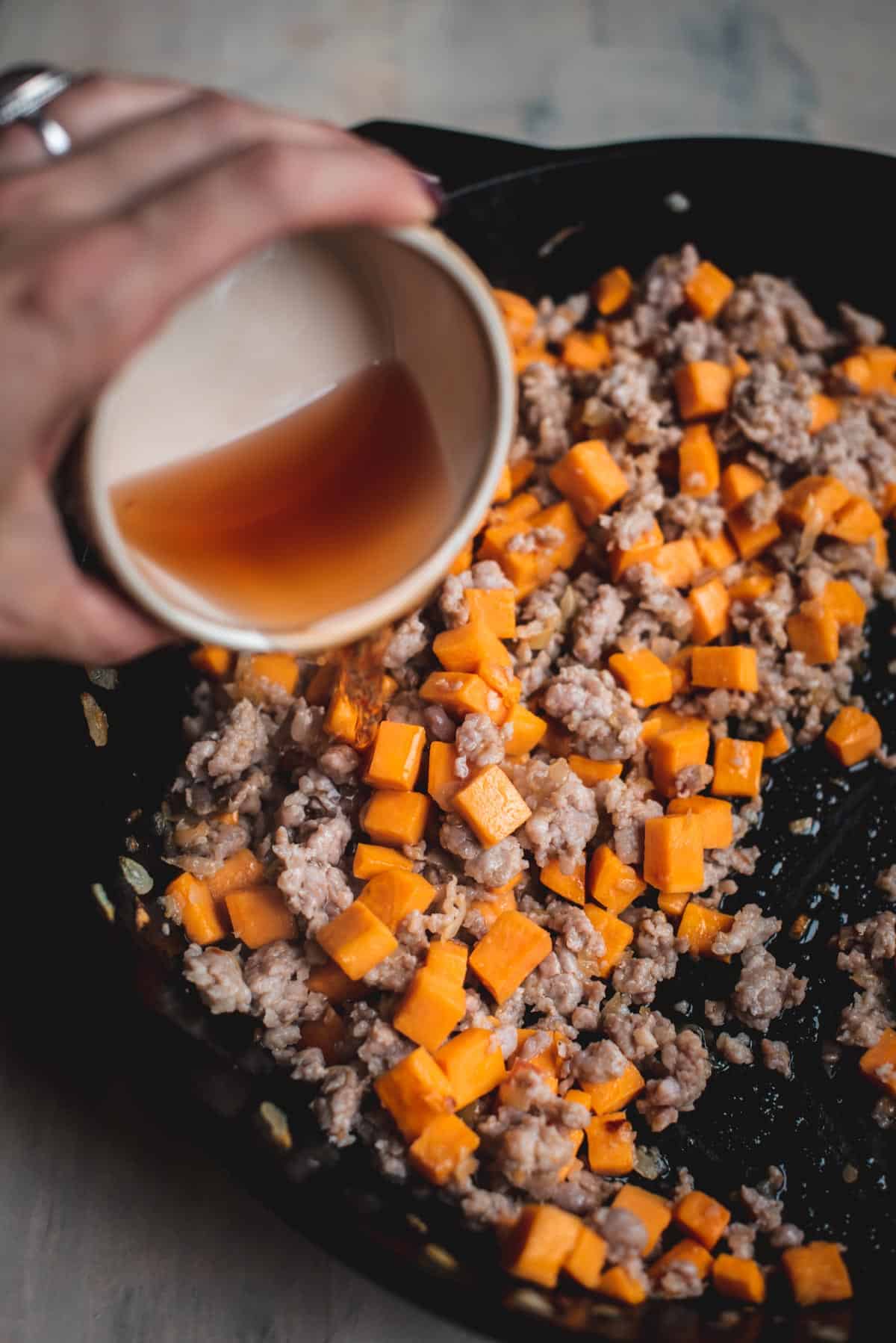 Sausage meat has been added to the potato, onion and gsrlicf mixture, in a black skillet. A hand is pouring in chicken stock to deglaze the pan.