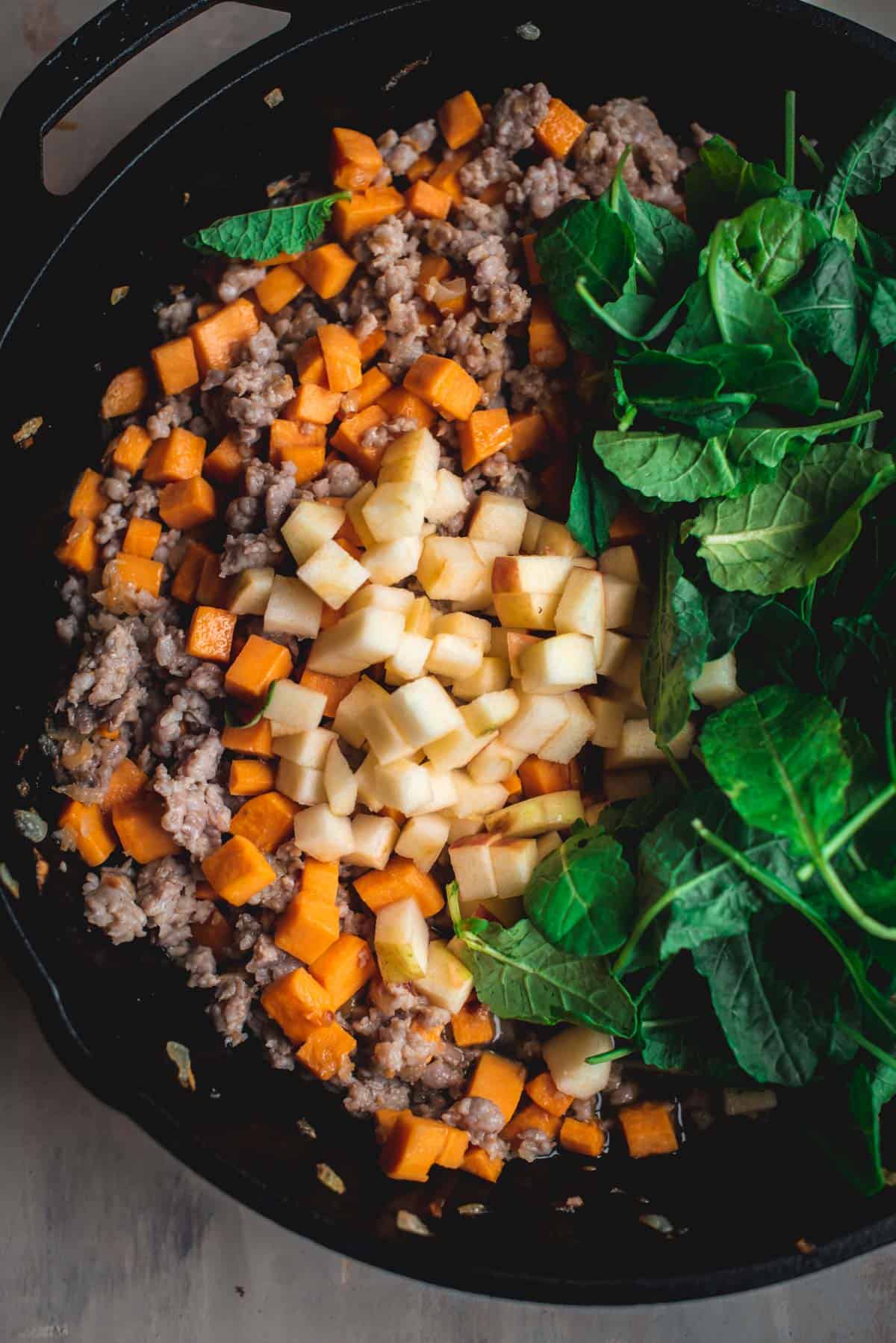 The skillet containing the sausage and potato stuffing has now been topped with apple chunk and green leave like spinach and kale. The ingredients are sitting on the top of the skillet and have not yet been incorporated. 