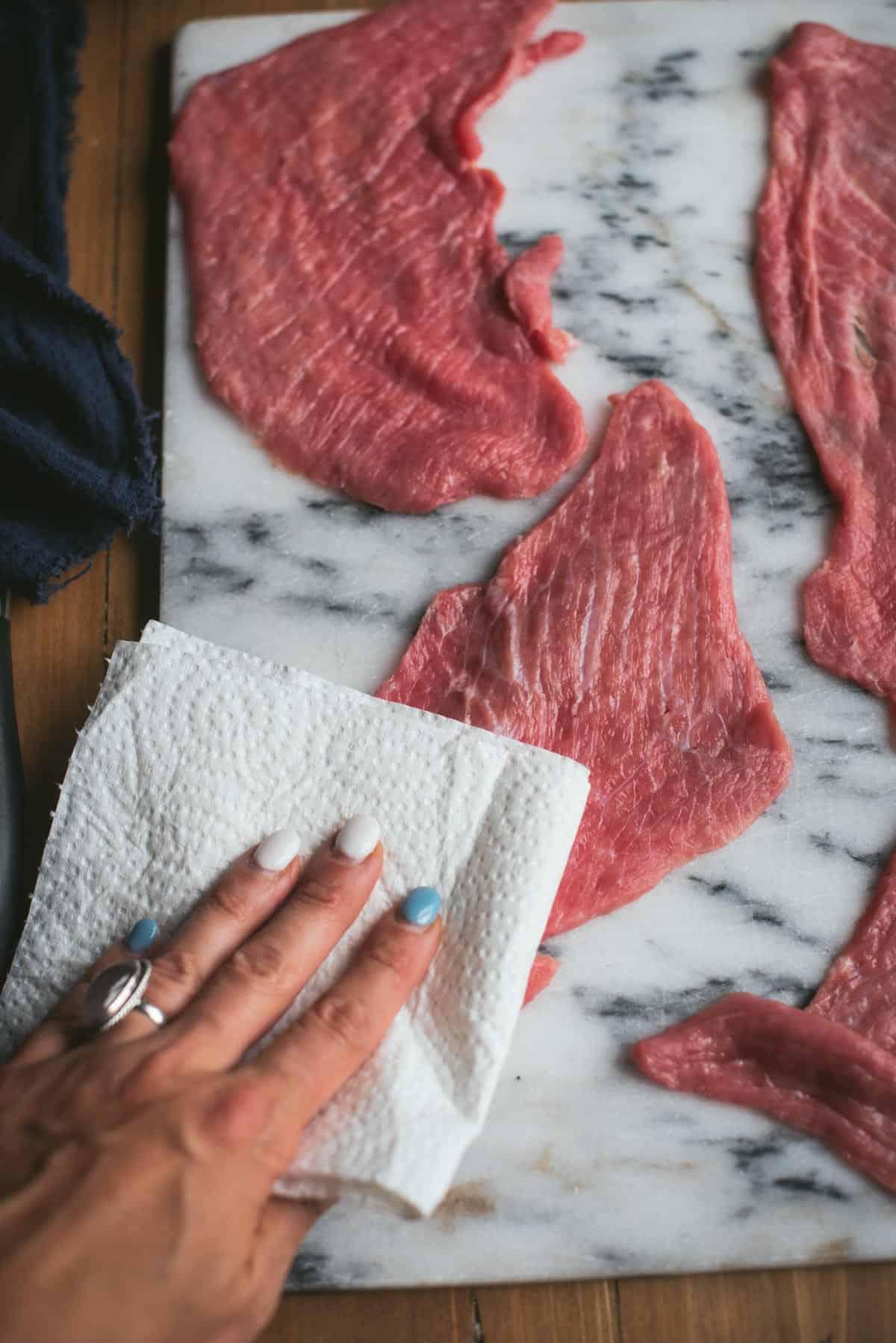 4 pieces of veal cutlet are laying on a white marbled chopping board and a hand is drying up any excess moisture with a piece of kitchen towel.