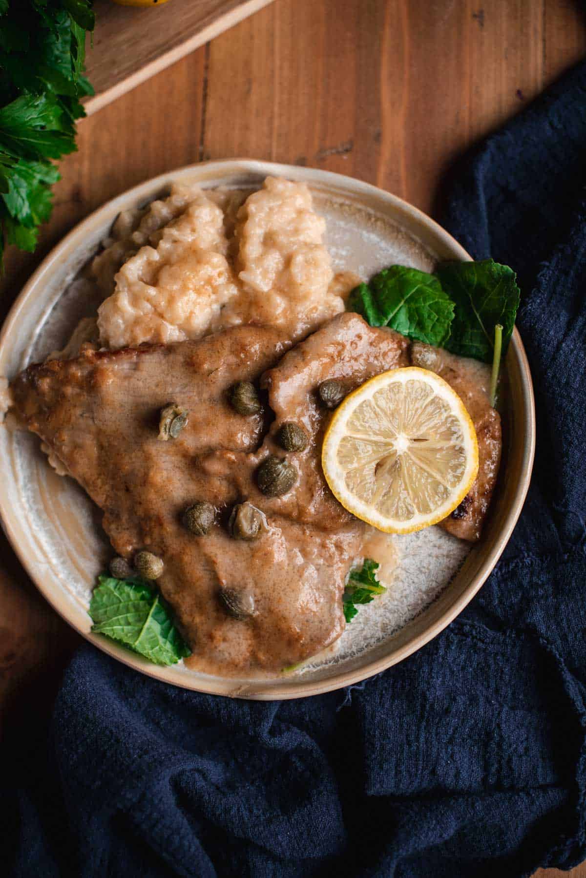 Veal Scallopini is placed on a cream colored ceramic plate, on a wooden table. The veal cutlets are coated in the buttery sauce and it is served with a side of parmesan risotto, some salad leaves and a lemon ring.