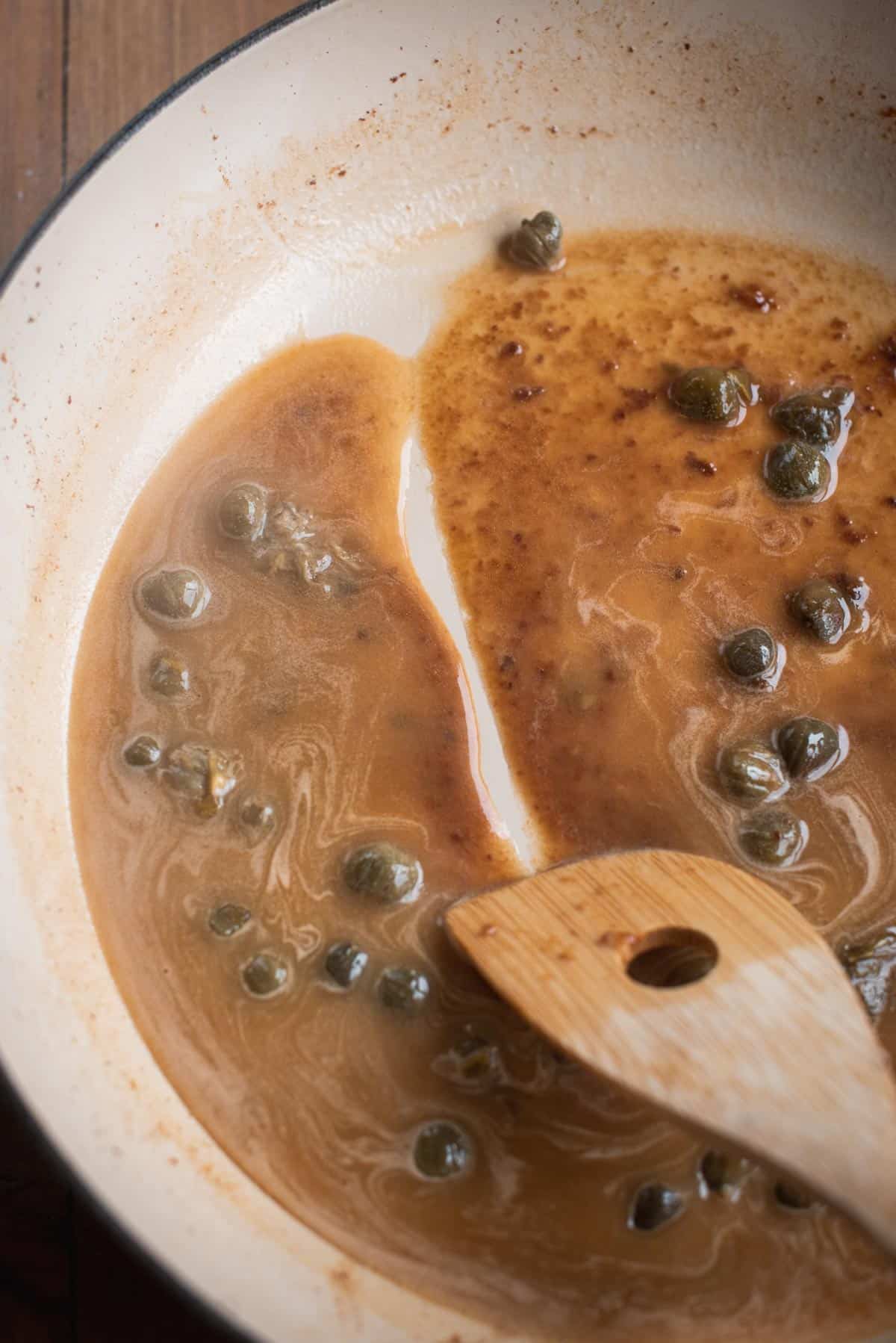A sauce is made in a large white saucepan, a wooden spoon is being used to stir all the ingredients together and scrape and tasty drippings of the bottom of the pan.