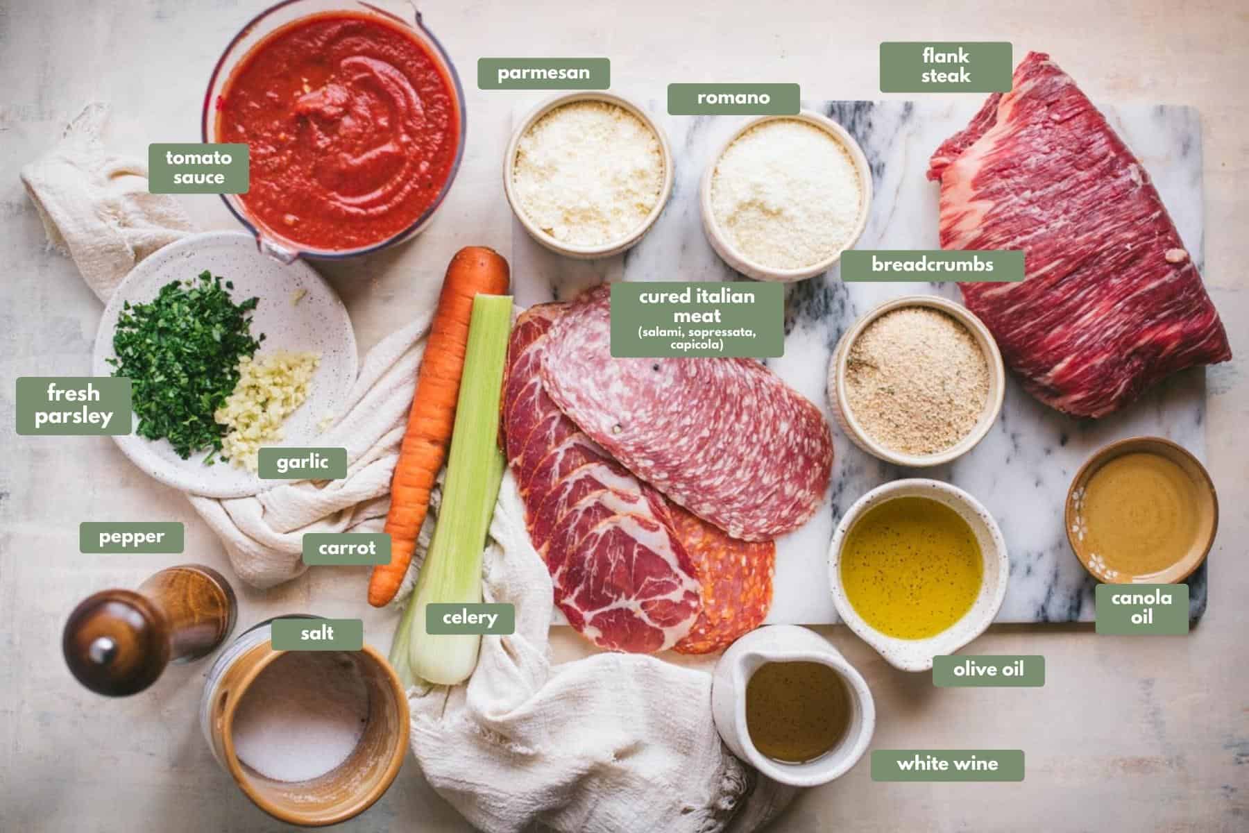 All of the ingredients for this beef braciole. There is flank steak sitting on a white chopping board with a selection of cured meats and a large celery and large carrot. There are also ramekin dishes filled with parmesan and romano cheese, breadcrumbs, olive oil  and canola oil. 
On the table there is a small jug of white wine, salt and pepper, a plate with fresh parsley and fresh chopped garlic on a white terrazzo plate, and a large jug of tomato sauce.