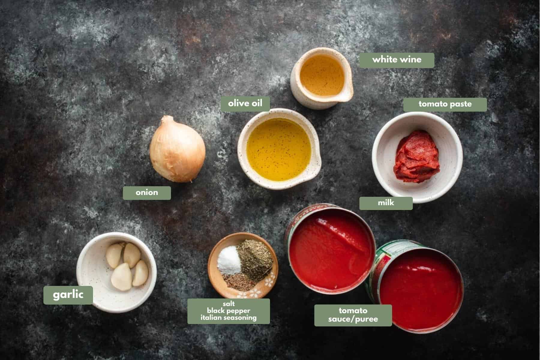 All the ingredients for this pasta sauce recipe, separated into individual containers sitting on a slate grey work bench. 
There is a small white jug with white wine, a small white ramekin dish containing olive oil, another white ramekin dish with a small amount of tomato paste, 2 can of tomato sauce, a brown dish containing salt, black pepper and Italian seasoning, a white dish with 4 cloves of garlic and 1 brown onion.