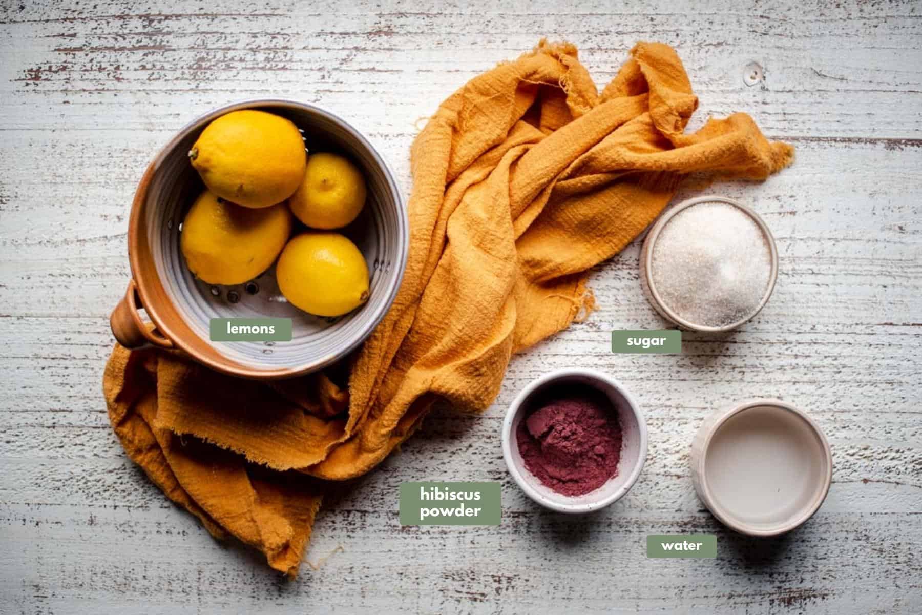 overhead image of ingredients for hibiscus lemonade.  Lemons are sitting in a gold and white ceramic bowl over a yellow linen napkin.  Sugar, water, and hibiscus powder in cream colored ramekins.