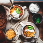 2 white bowls of pumpkin beef chili sitting on a tablecloth on a wooden tray. The chili is topped with cheese, sour cream and jalapenos. There is a bowl of grated cheese on the tray, also sour cream and a pot of jalapenos. Part of the pumpkin shaped pot with the chili inside can be seen also.