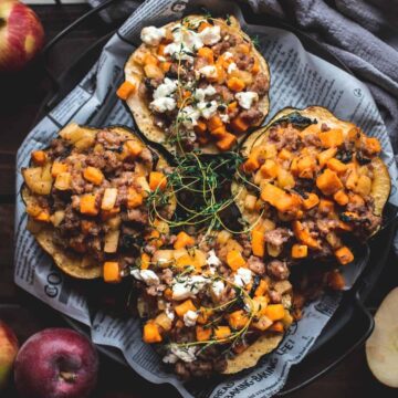 4 half acorn squash stuffed with bright orange ingredients like spiced sausage meat and sweet potato. They are sitting on a newspaper lined plate and have been garnished with some cheese and herbs.