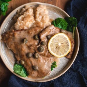 Veal Scallopini is placed on a cream colored ceramic plate, on a wooden table. The veal cutlets are coated in the buttery sauce and it is served with a side of parmesan risotto, some salad leaves and a lemon ring.