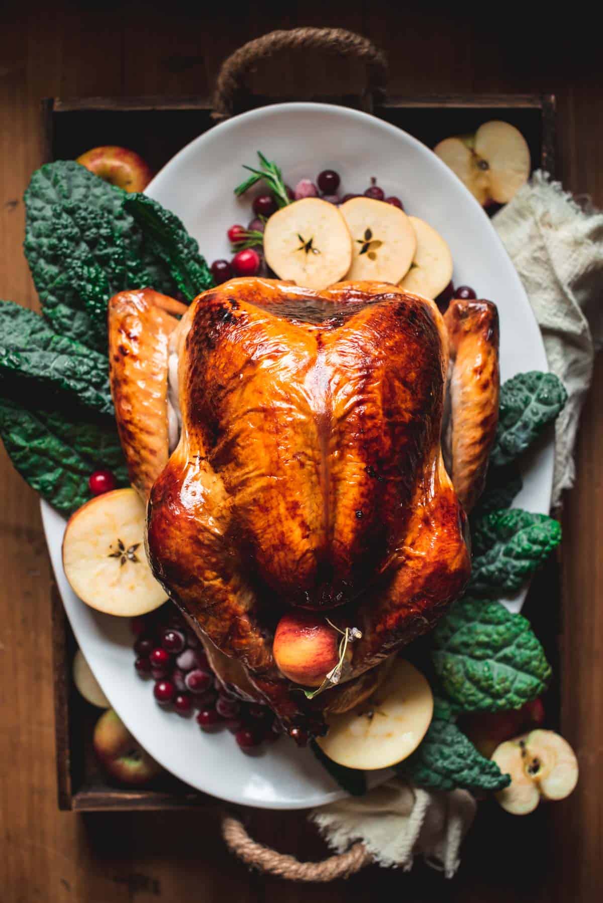 Brined and perfectly roasted turkey with brown crispy skin and sitting on a white platter on top of salad leaves, apples and cranberries.