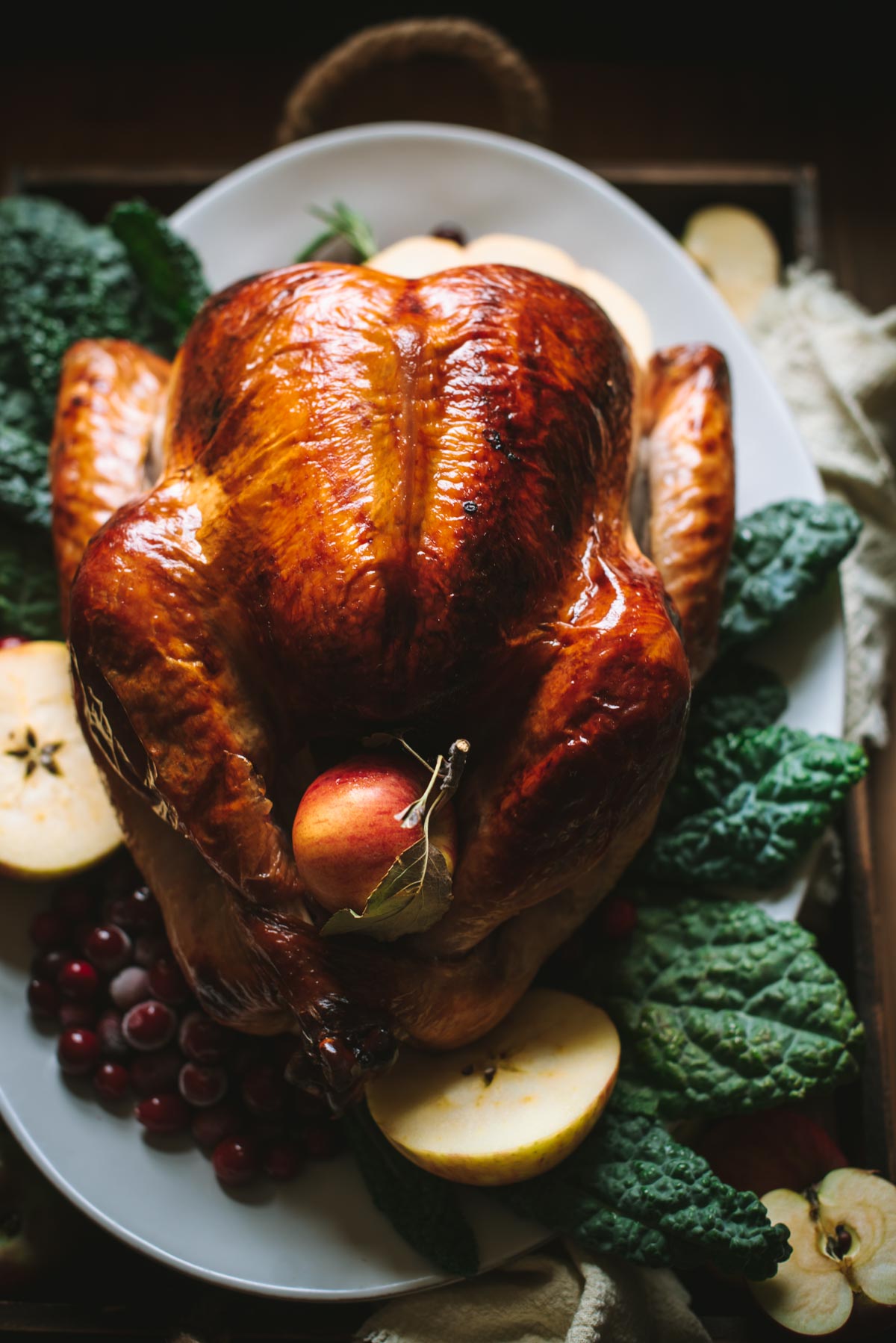 Close up of crispy baked turkey. It has been brined in apple cider and placed on a white serving platter. It has been decorated with salad leaves underneath and slices of apple scattered around, There is also a full apple sitting where the legs cross.