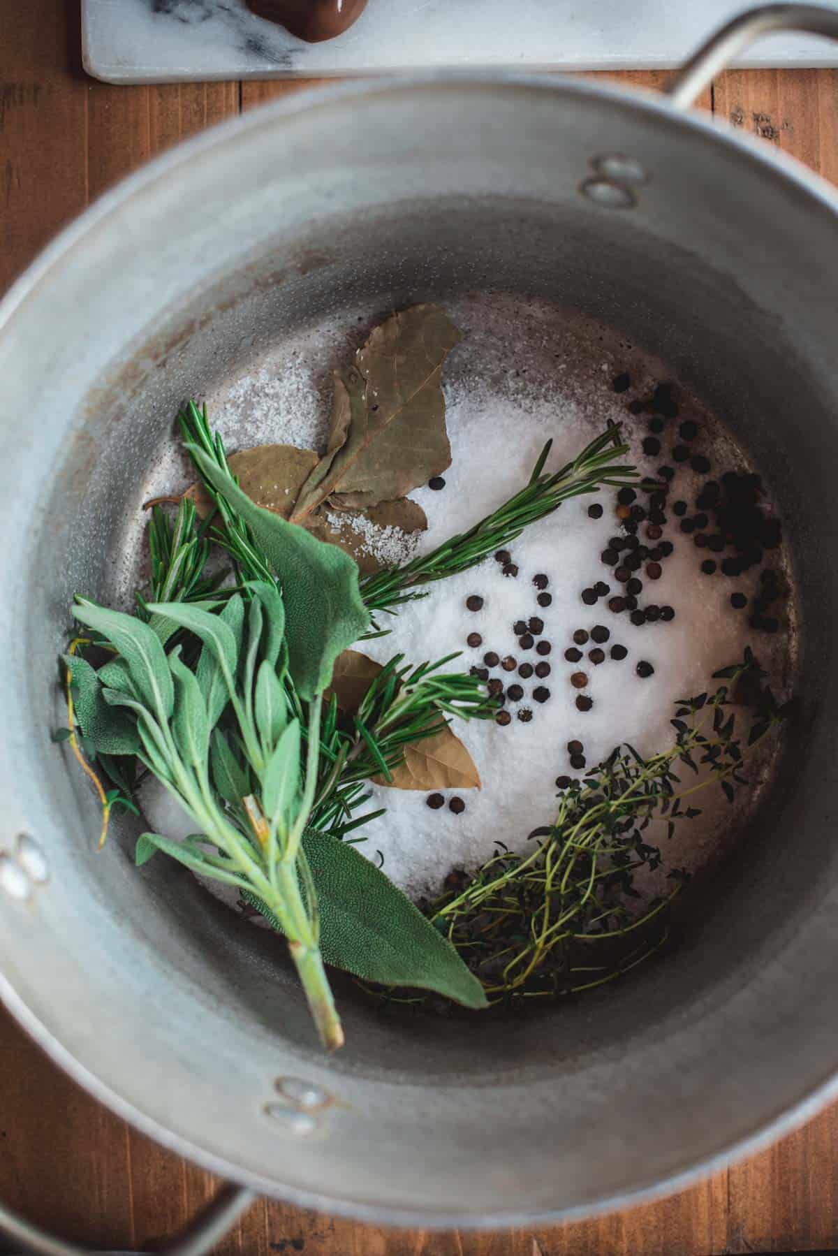 In the bottom of a large silver stock pot there is rosemary, thyme, sage, bay leaves, peppercorns, and salt.