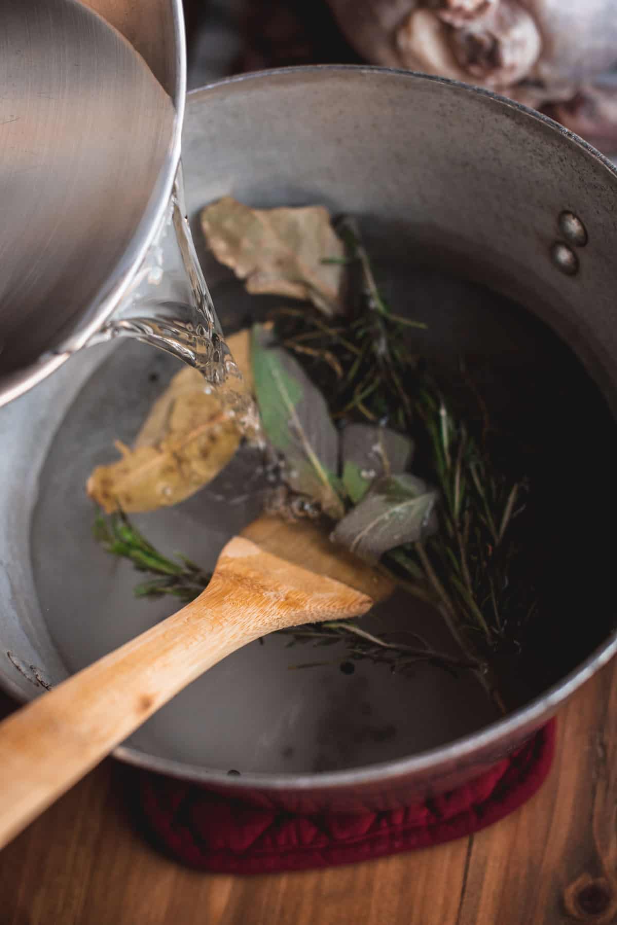 Someone is pouring water into the silver stock pot with the herbs. A wooden spoon is being used to stir them.