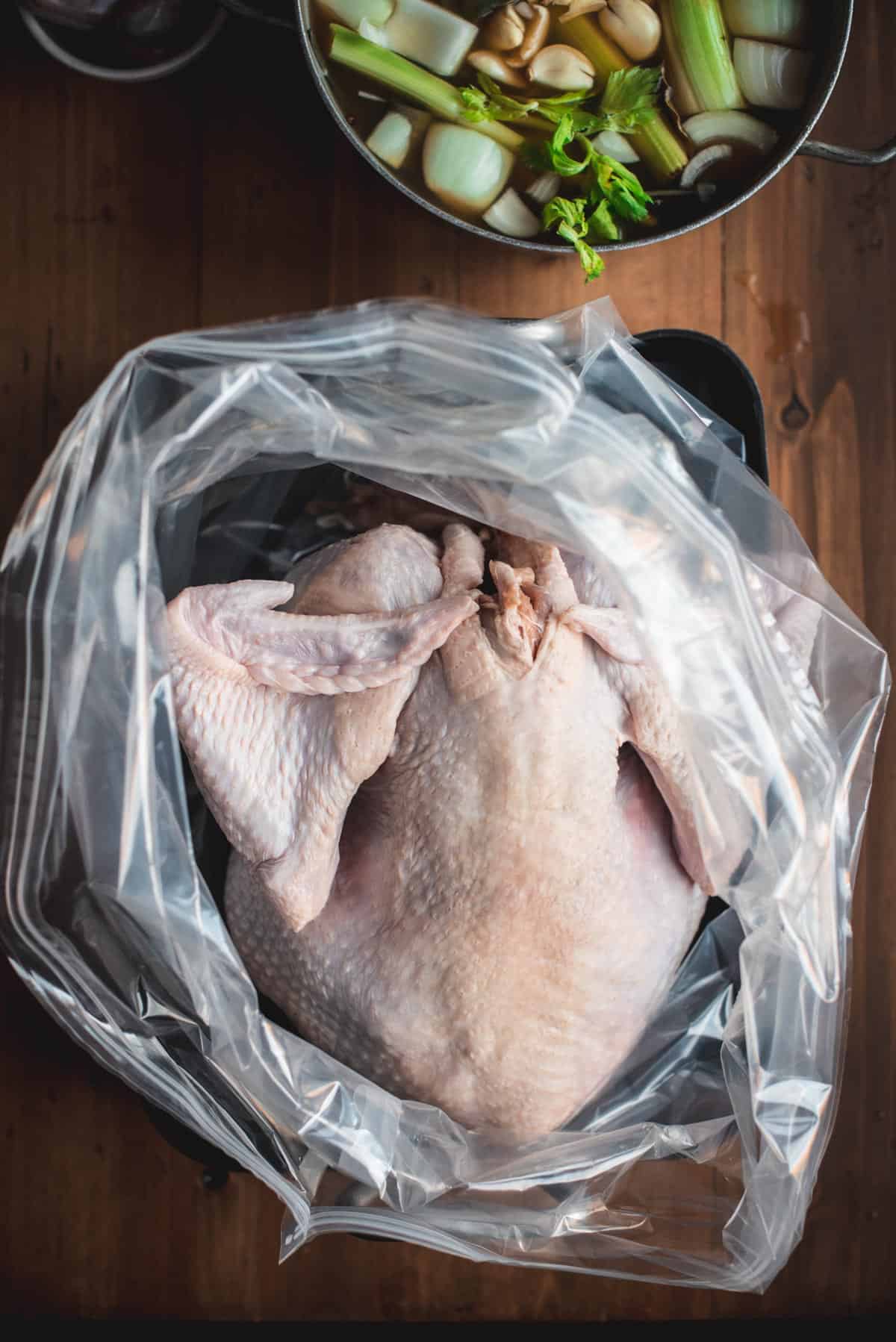 Full raw turkey sitting in a clear brining bag in a baking pan. The bag is open but there is nothing other than the turkey inside it.
Above the turkey there is a pot with liquid, herbs and onions and garlic inside it.