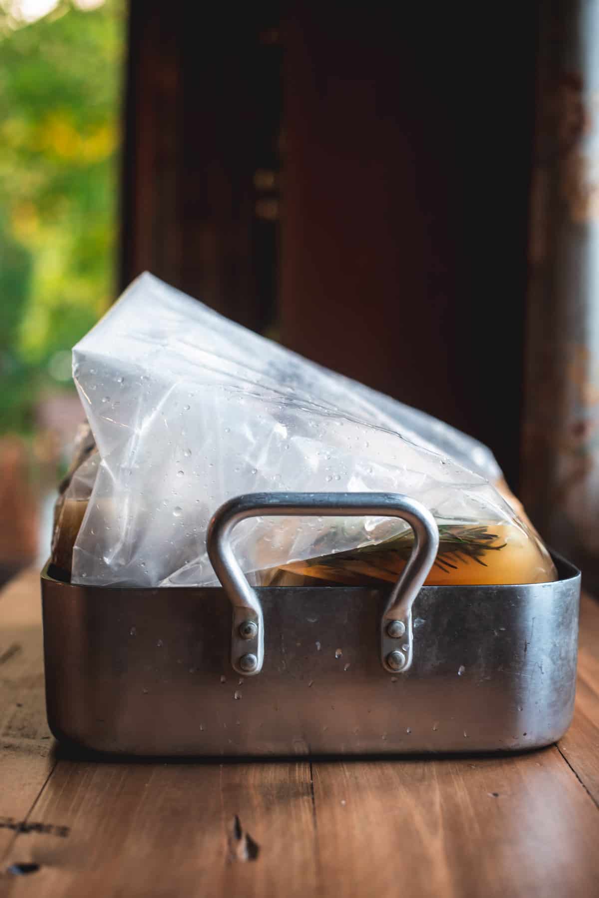 The brining bag is sealed with the liquid and herbs inside. it is sitting in a deep baking pan on a wooden table.