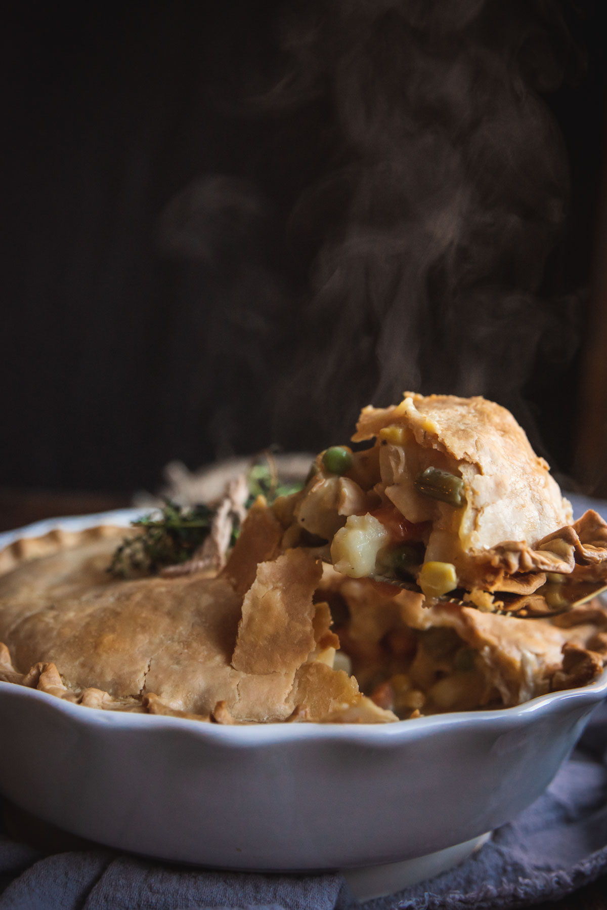 The creamy chicken pot pie is in the white pie dish. A piece is being lifted out, but it has not yet cleared the pie dish. The background to the photo is black and you can see the steam billowing out of the pie as it has been cut.