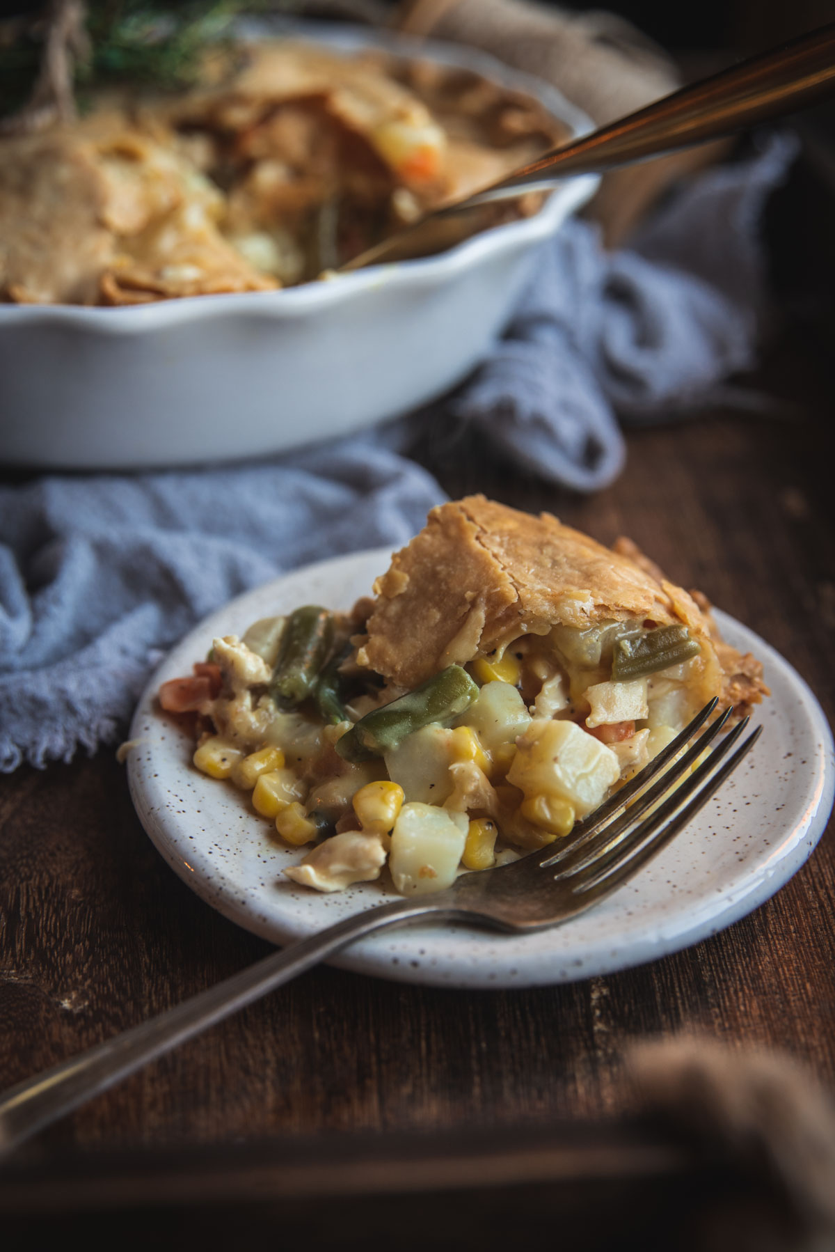 Up close picture of slice of chicken pot pie. The piece of pie is sitting on a white terrazzo plate with a sliver fork. You can see its filled with veg vegetables like sweetcorn, green beans, hash browns and carrots. In the background, out of focus, is the white pie dish with the remains of the pie inside.