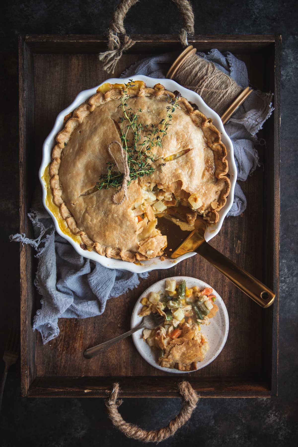 View from above of creamy chicken pot pie in a white pastry dish. A portion has been cut out with a gold serving knife and placed on a white plate. The serving knife is sitting in the whole made by the section of missing pie. The pie and the plate are sitting on a wooden tray and the is a blue dish cloth there aslo.