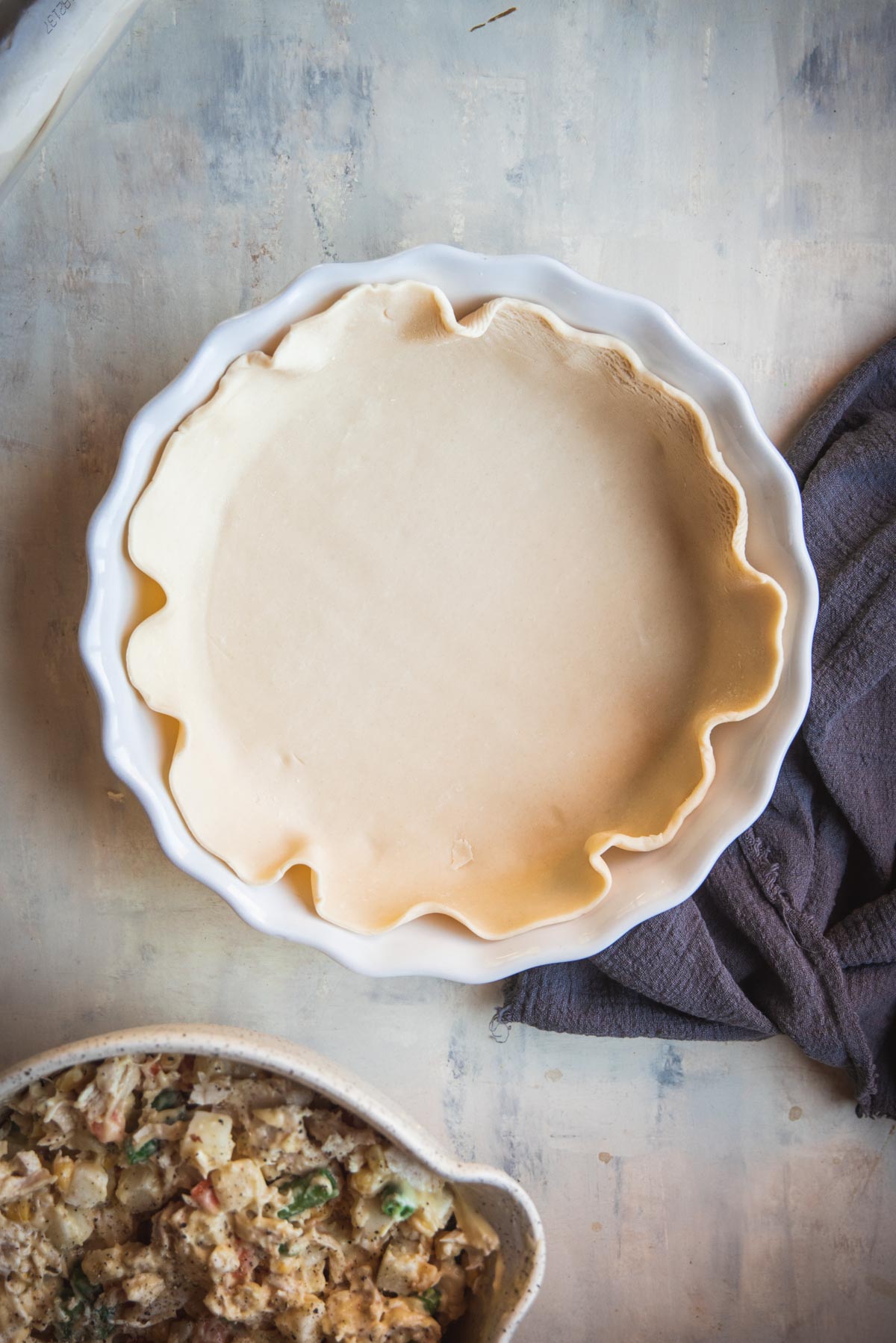 A white pie dish has been lined with uncooked pastry. There is another bowl in the bottom left corner containing the pie filling.