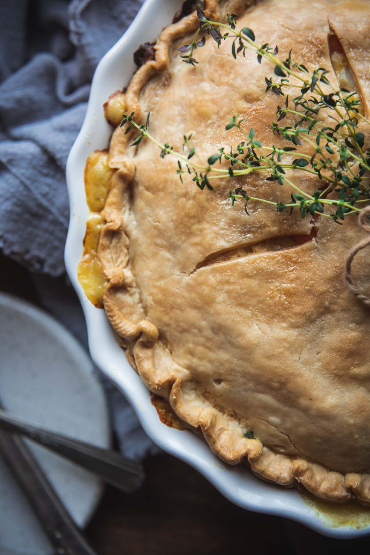 Partial view of the pot pie up close. It is in a white pie dish and the pastry on top is golden brown, It has also been garnished with some herbs.