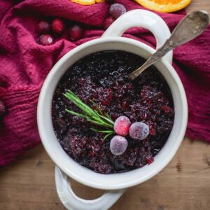 Cranberry Sauce with Grand Marnier in a white bowl with handles. It is sitting on a wooden table draped in red fabric. The sauce is topped with frosted cranberries and some fresh herbs. There are orange slices and extra cranberries scattered on the table.