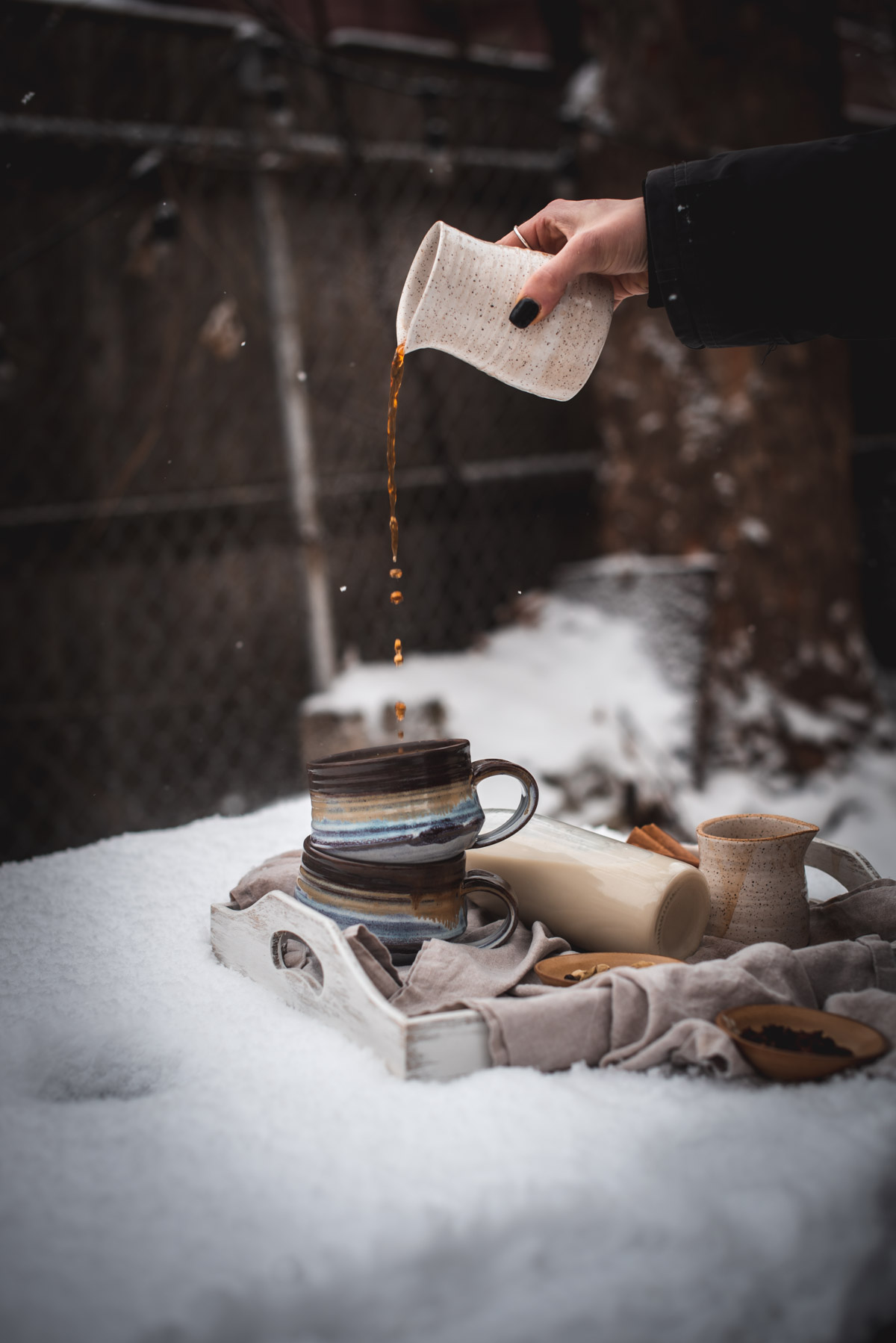 Two ceramic mugs in layered colors of blue, lavender, and brown stacked on top of each other on a white beverage tray.  Chai tea latte syrup is pouring into the top mug from a white ceramic pourer.  The beverage tray is set on a snow covered table.