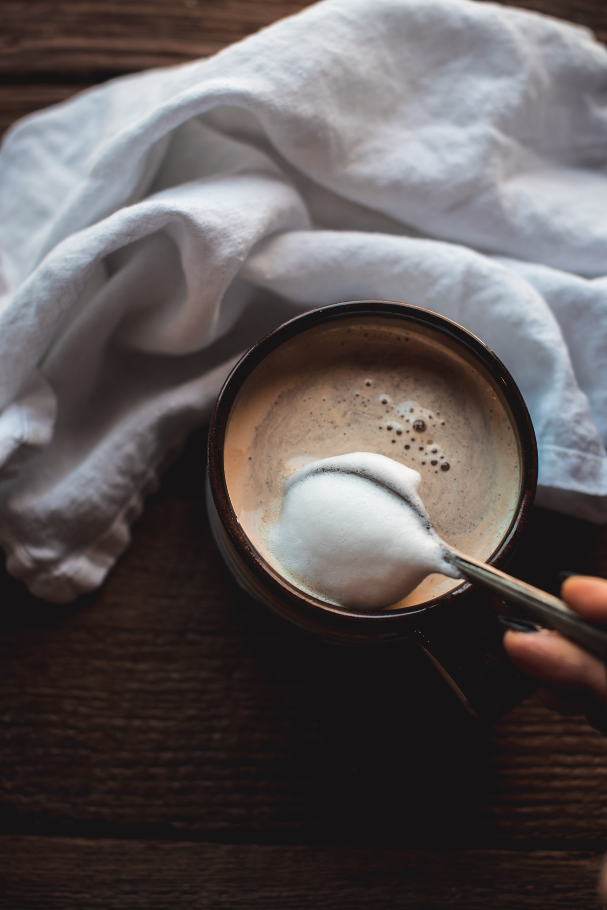 Close up image of spoon ladling milk foam on top of a latte inside a brown mug.  The mug is set on top of a wooden background with a white linen napkin.