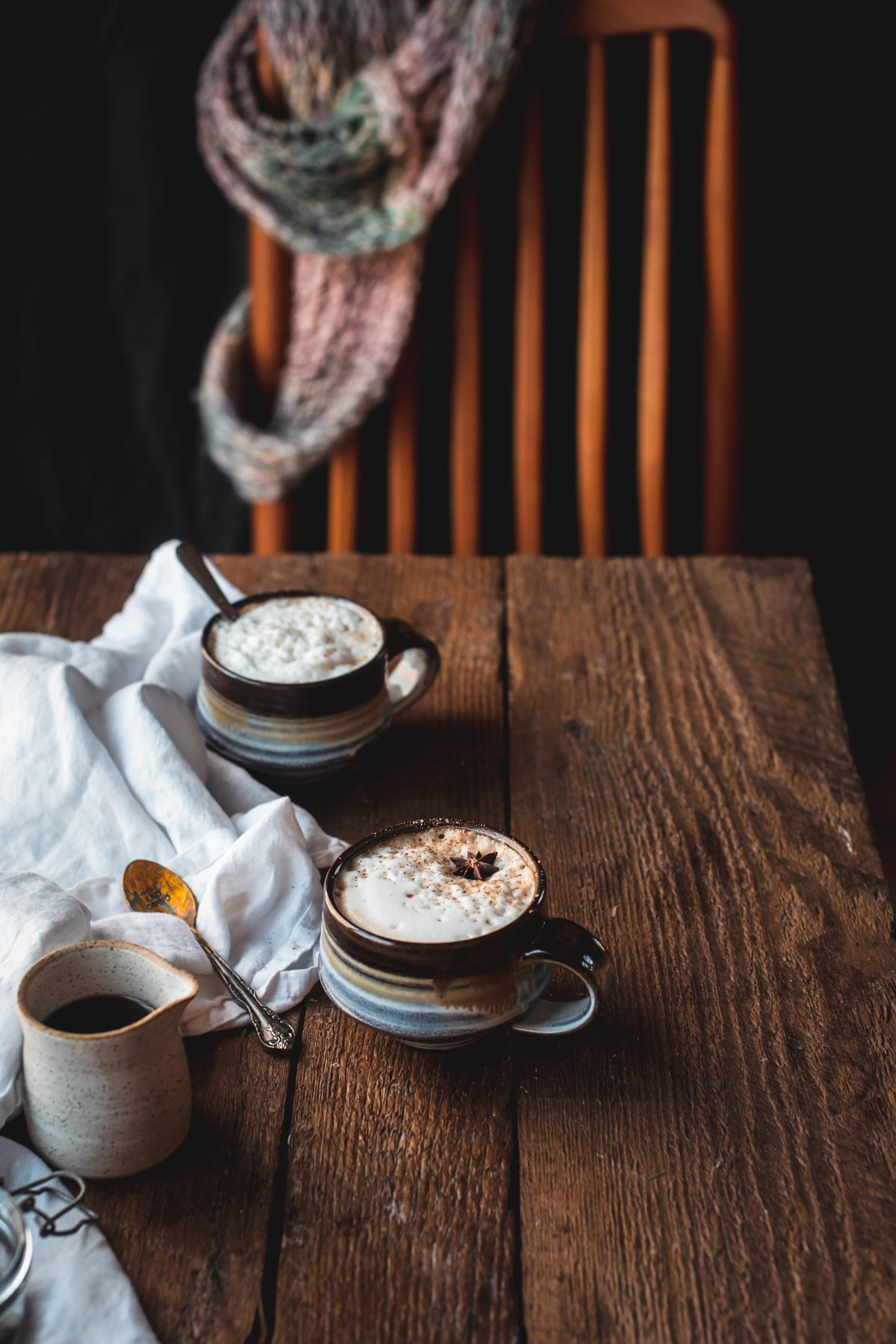 Two coffee mugs filled with foamy milk and sprinkled with cinnamon on top of a rustic wood surface.  A ceramic carafe and antique spoon are next to the mugs on a white linen napkin.  A wooden chair draped with a scarf is in the background.