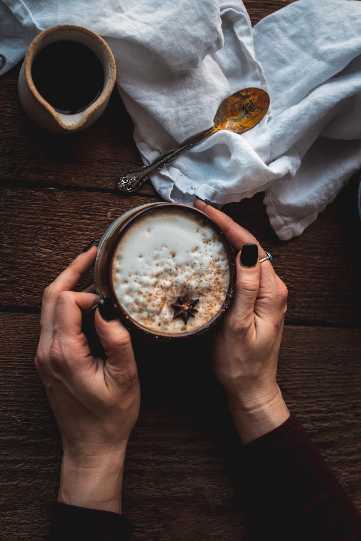 Two hands are holding a brown mug filled with dirty chai latte.  The top of the latte is sprinkled with spiced and topped with anise.  The background is a rough wooden surface with a linen napkin, antique spoon, and ceramic pourer.