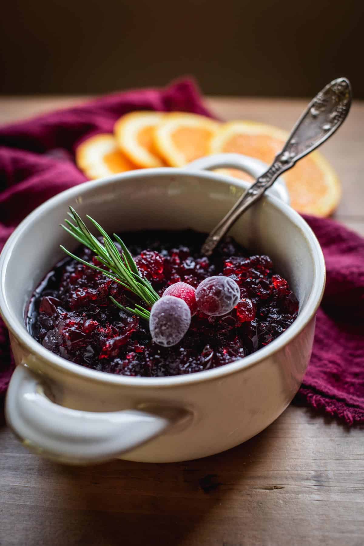 Cranberry Sauce in a white bowl with handles. It is sitting on a wooden table draped in red fabric.  The sauce is topped with frosted cranberries and herbs. There is orange segments lying on the red fabric on the table.