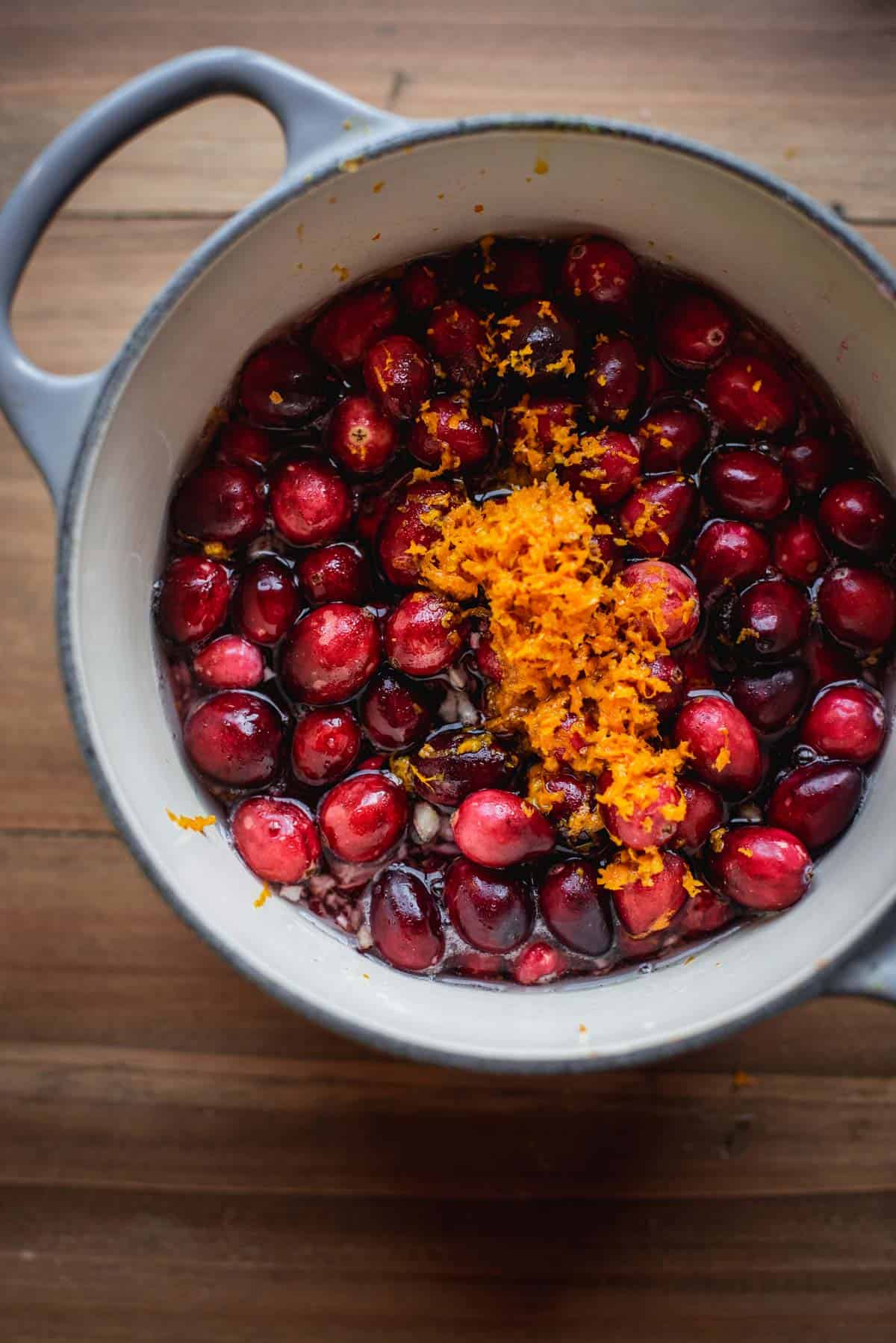 A large pot sits on a wooden table and is filled with full cranberries, orange zest, blended cranberries, grand marnier and water.