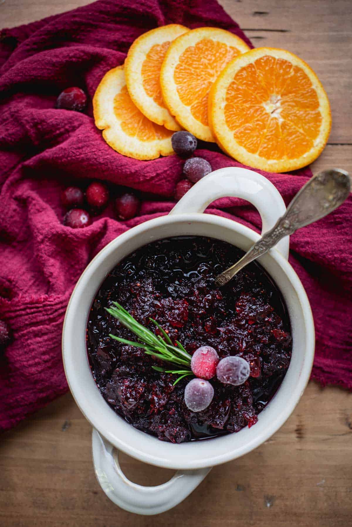 Cranberry Sauce with Grand Marnier in a white bowl with handles. It is sitting on a wooden table draped in red fabric. The sauce is topped with frosted cranberries and some fresh herbs. There are orange slices and extra cranberries scattered on the table.