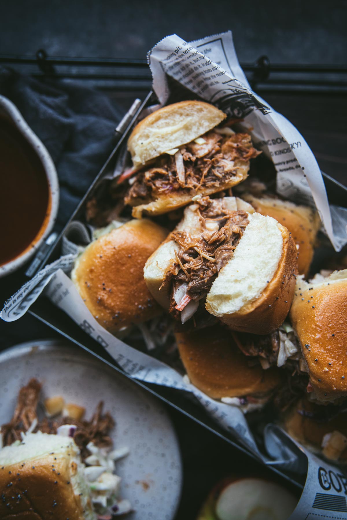 Pulled pork sliders sitting in a box lined with newspaper. Some buns are on their side so you can see all the meat and juices inside the bun. There looks like some kind of dipping sauce next to the box and a small white terrazzo plate with 1 slider on it. It is very full and the contents has managed to fall out if the bun a bit. 