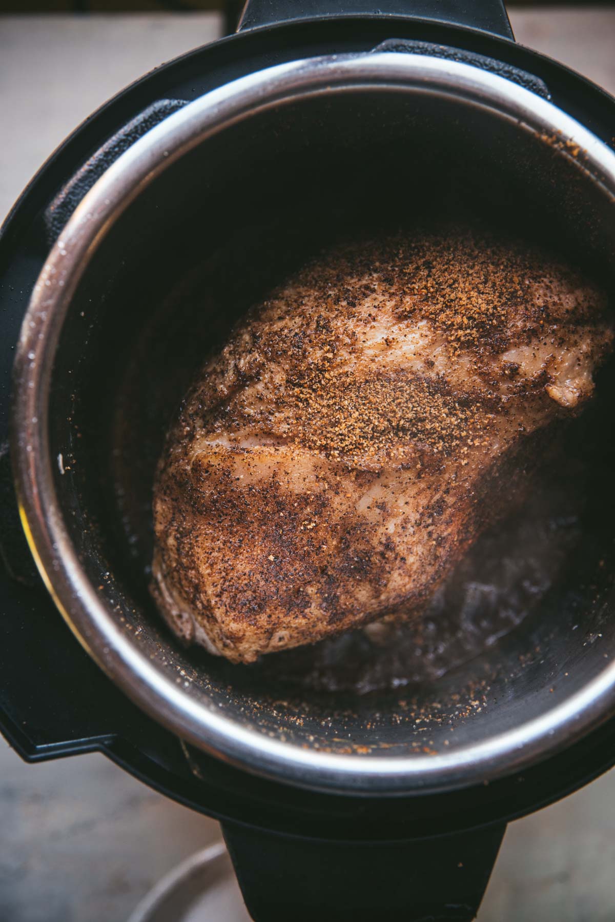 The pork is inside the black slow cooker cooking. The pork is still whole and has not yet been shredded.