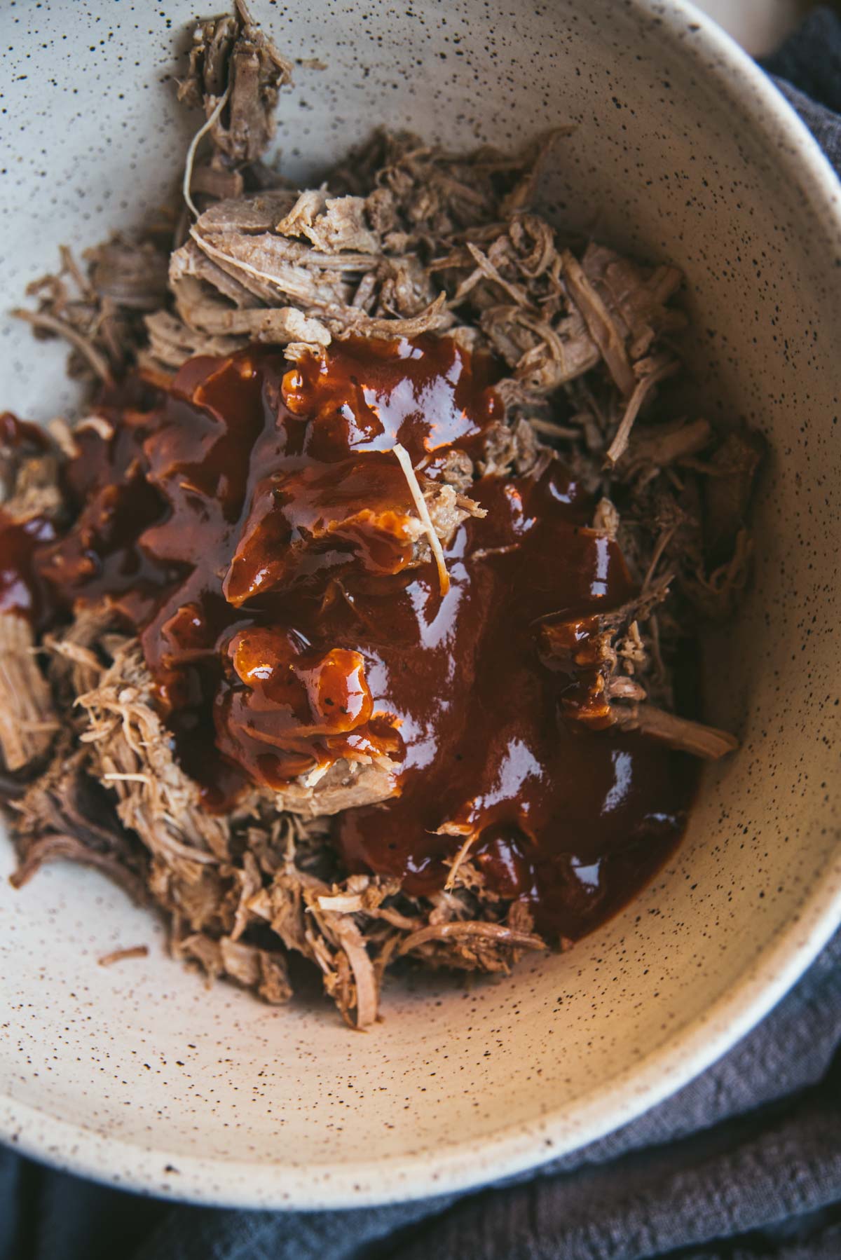 The pork has been placed into a deep terrazzo bowl and has been shredded, it has been topped with plenty of bbq sauce but it has not yet been mixed together.