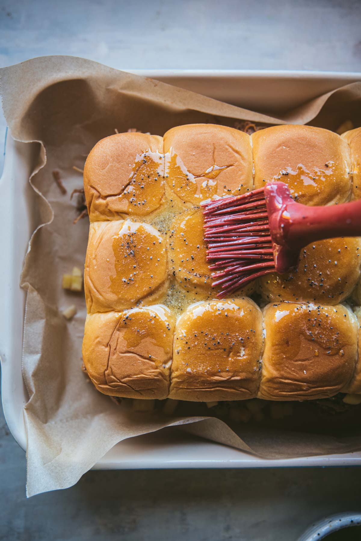 The sheet of burger buns have been placed in a tray covered with baking paper. A red silicone brush is brushing melted butter and poppy seeds on top of the buns. 