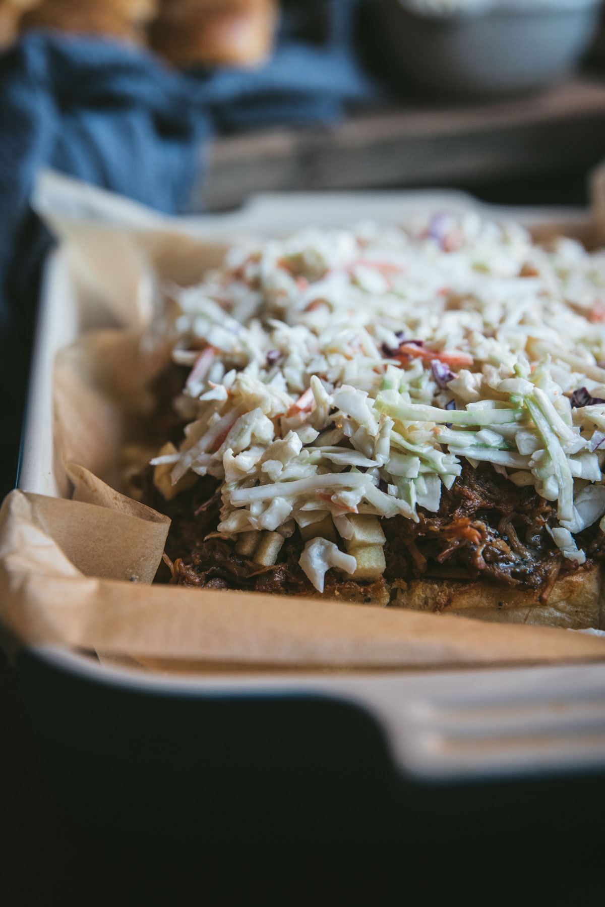 The top layer of buns is missing. The bottom layer remains on the covered baking tray and the pulled pork is placed on top of the buns to cover. Coleslaw has been placed on top of the pork and spread to cover evenly.