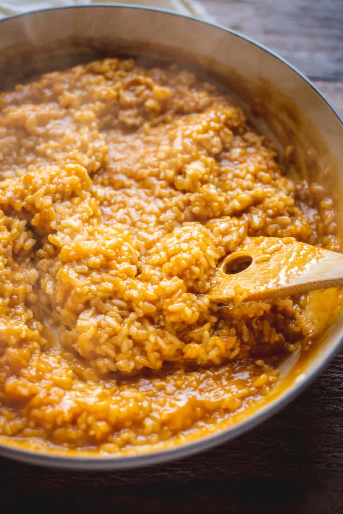 Orange color risotto without too much liquid. A wooden spoon with a hole through it is slowly moving the risotto around the pot.