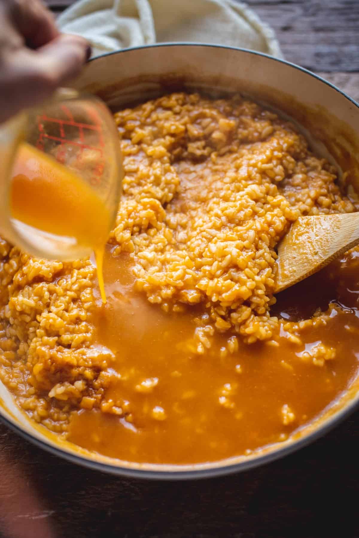 A jug containing more stock and pumpkin is being poured into the pot holding the risotto. The wooden spoon is inside the mixture to stir in the liquid.