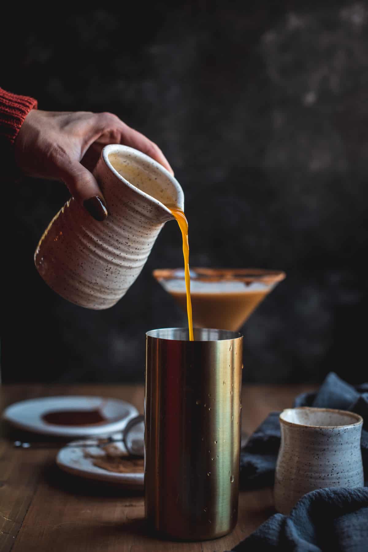 An amber color liquid is being poured from a white ceramic jug into the cocktail shaker. There is a finished cocktail in the background and the plates used to rim each glass.