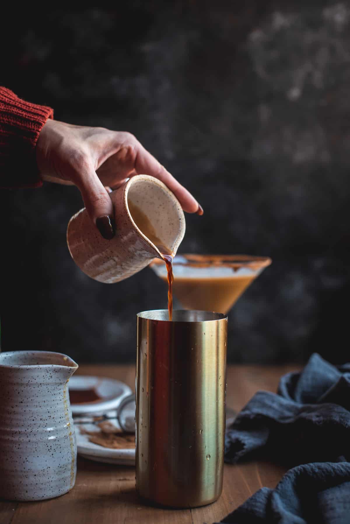 Espresso is being poured into a metal cocktail shaker base. There are some other ingredients laying around this image like a white ceramic jug and material. There is also a finished cocktail in the background.