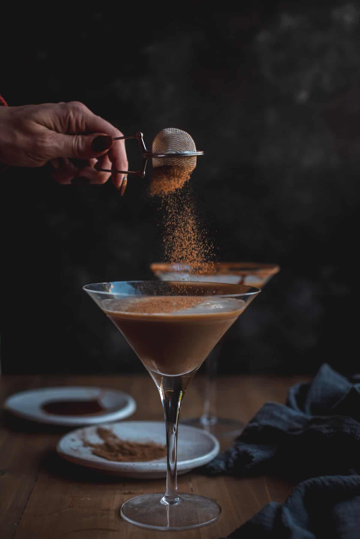 2 pumpkin espresso martinis sitting next to each other on a wooden table, 1 is behind the other so barley visible.  Cinnamon is being sifted through a small strainer on top of the cocktail. There are 2 white plates on the table also, 1 has cinnamon powder and the other is a sticky syrup so that the martini glass rims can be decorated with cinnamon powder.