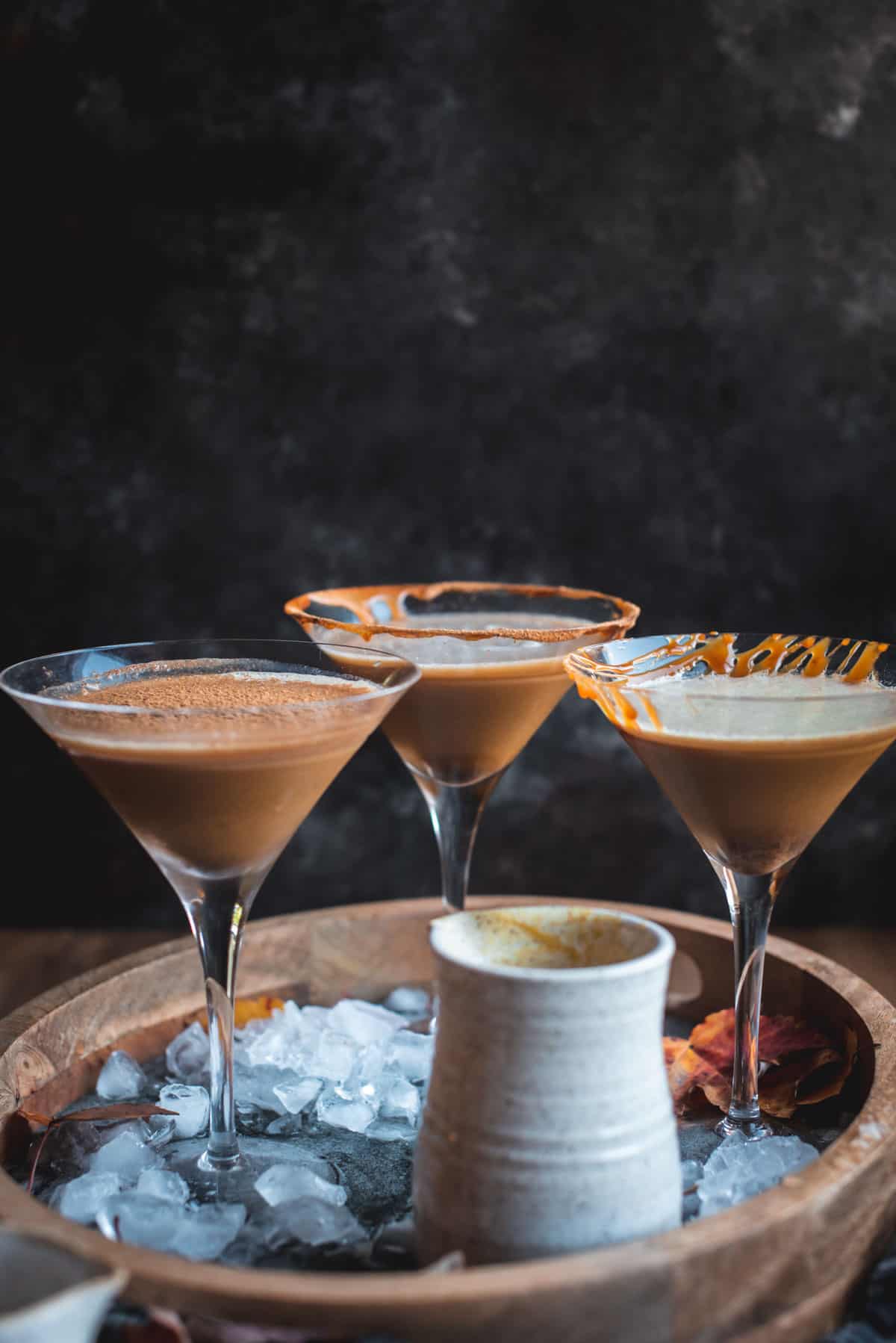 3 Pumpkin Spice Latte Martinis are in long stem martini glasses on a wooden tray next to ice cubes. 2 of the cocktails have been topped with a drizzle of caramel and 1 has been topped with some cinnamon powder. There is a small white ceramic jug on the tray too, contents can not be seen.