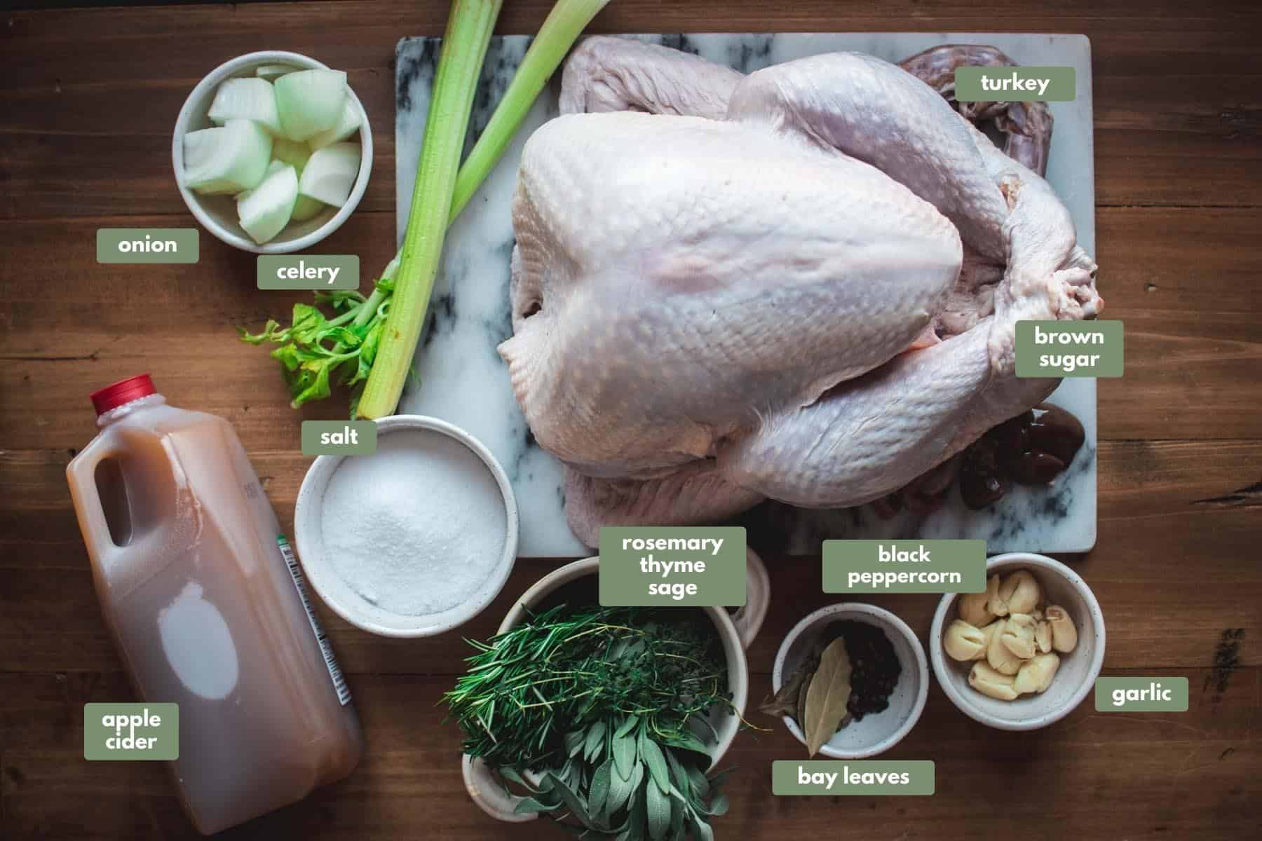 Ingredients required to brine your turkey. There is a turkey sitting on a white marble chopping board next to some celery. There is also bowls on the wooden countertop. One bowl contains garlic, another black peppercorns and bay leafs. In another bowl there is herbs like rosemary thyme and sage and also a bowl of salt. There is a bottle of apple cider and a bowl containing onion and celery.