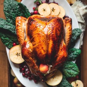 Brined and perfectly roasted turkey with brown crispy skin and sitting on a white platter on top of salad leaves, apples and cranberries.